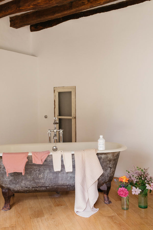 Minois bottle on the rim of a roll-top bath with pink towels draped over it and flowers in jars | Botā natural skincare.