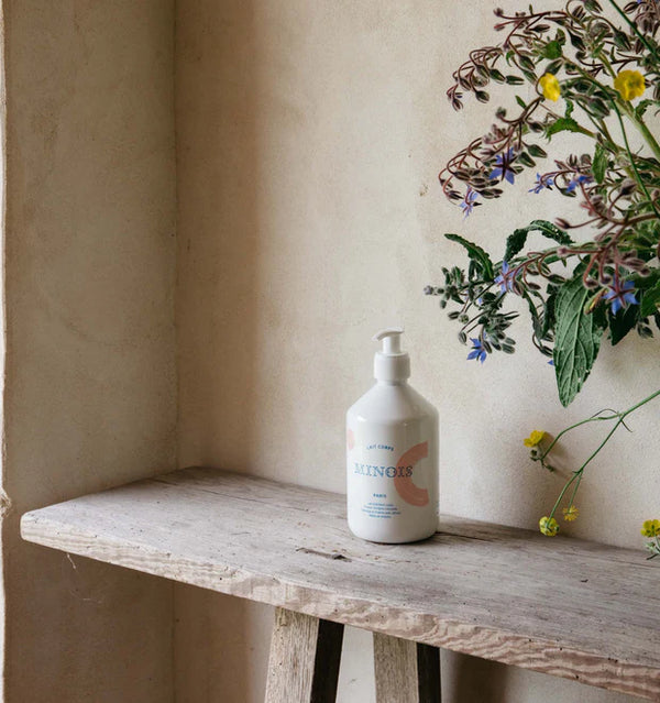 A single Minois pump bottle on a rustic wooden shelf beside wildflowers and a plaster wall | Botā natural skincare.