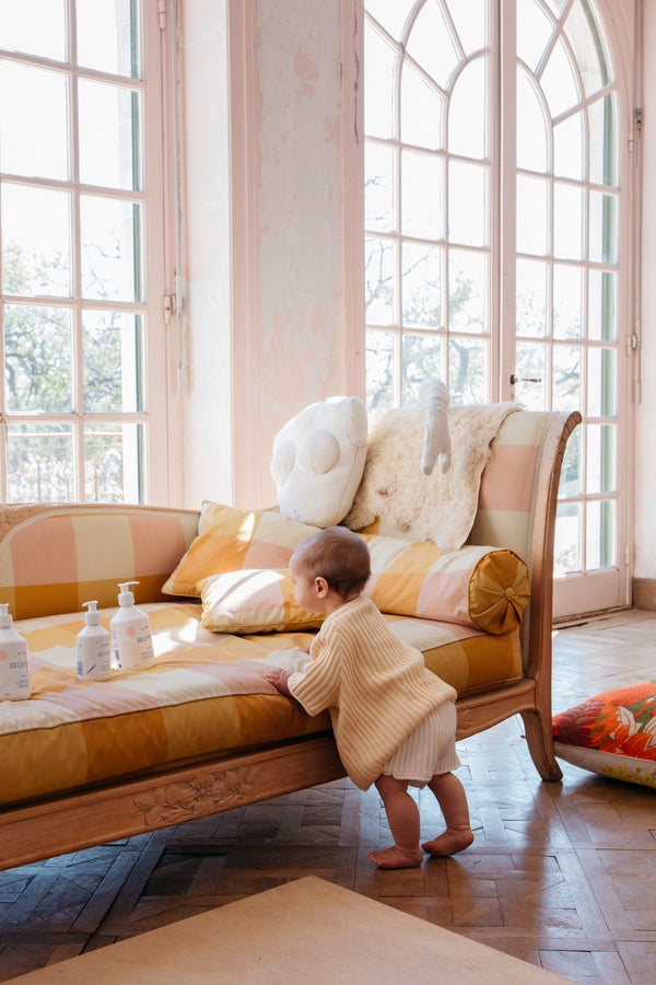 A child standing at a striped daybed with Minois pump bottles on the seat, sunlit room | Botā natural skincare.