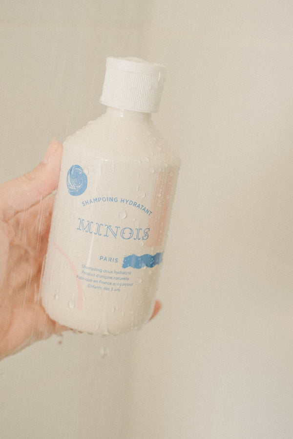 A hand holding a wet Minois shampoo bottle under running water in the shower | Botā natural skincare.